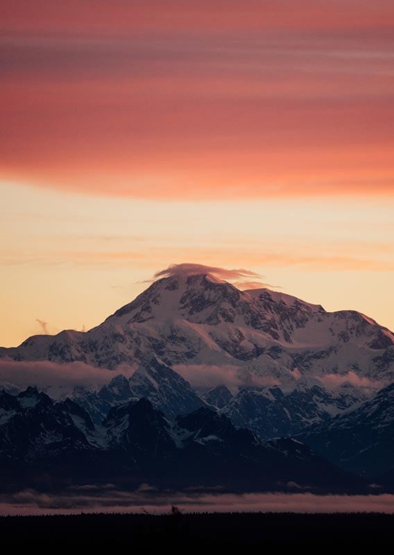 Denali, a tall mountain, rises above a later of clouds below a red sky.