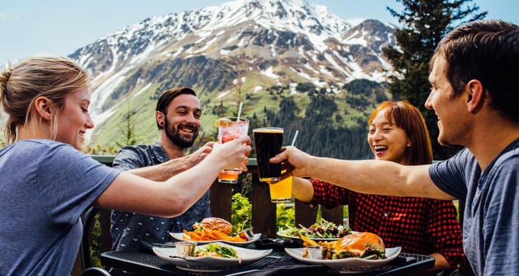 Four friends raise their glasses in cheers on a patio with mountains behind