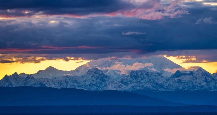 A view of Denali with glowing sunshine and dark clouds above.