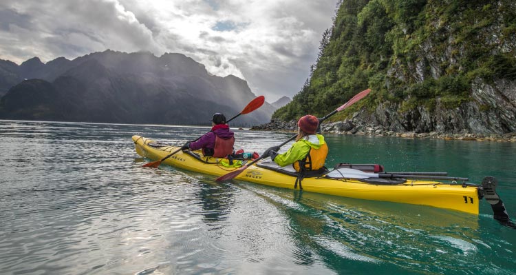 Two kayakers paddle around plant-covered, rocky cliffs.