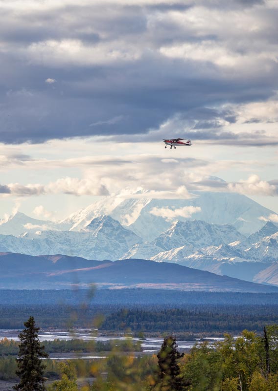 A plane flies above the tundra with Denali in the distance