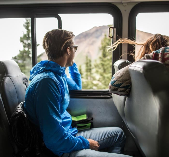 A man looks out the window of a bus driving through a forested wilderness