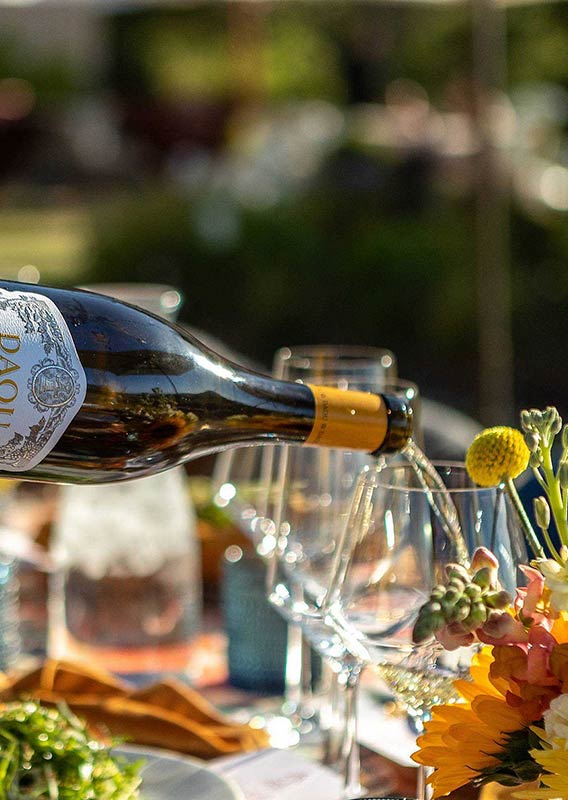A bottle of wine is poured on an outside table with flowers and food on it.