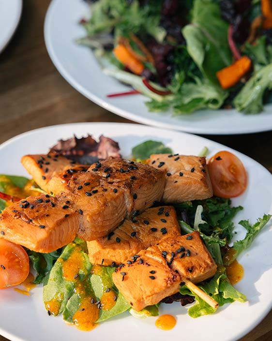 A close up image of salmon on a plate, with another plate of salad in the background.