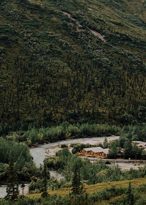 An aerial view of a series of wooden cabins alongside a river below a tree-covered hillside.