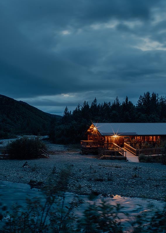 A wooden lodge near a river rushing below a forested hillside.