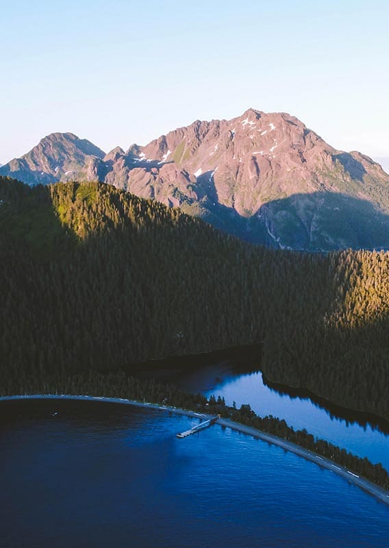 A dock stretches from a spit of land on a forested island among mountains