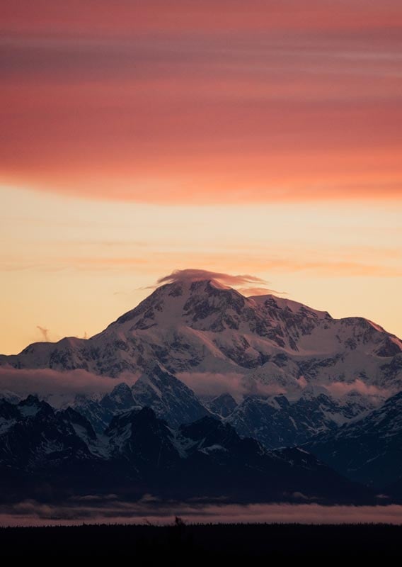 Denali, a large mountain rises high above clouds and below a red sky.