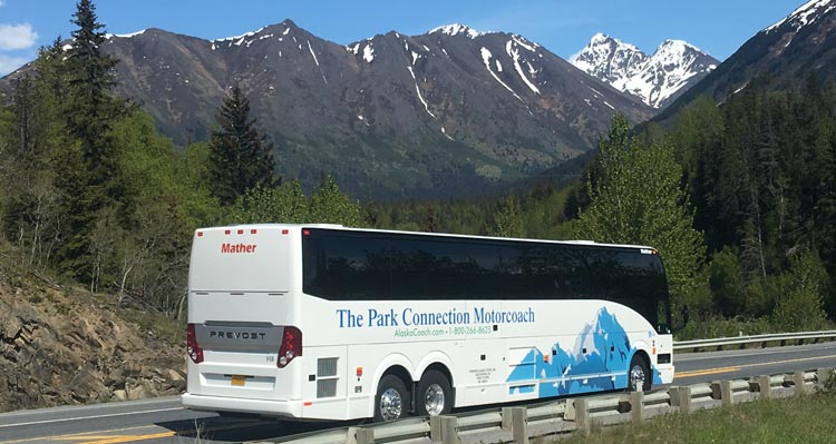 A motorcoach drives on a road beside lightly-snowy mountainsides.