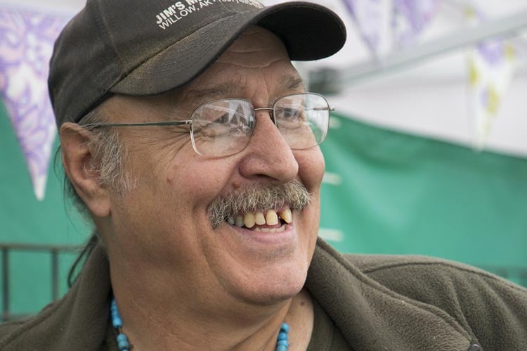 A man wearing a baseball cap glasses and a blue necklace smiles