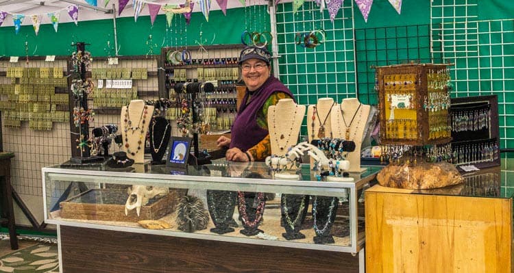 Mary-Ann stands in a market tent showcasing many displays of jewelry