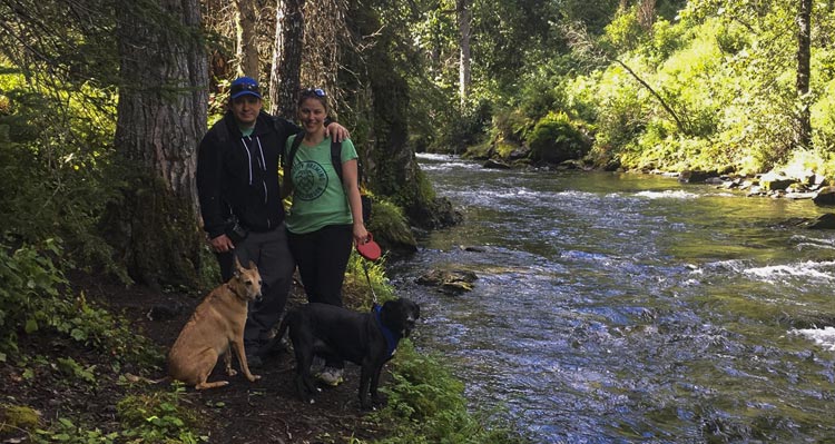 Two people and a dog stand alongside a river.