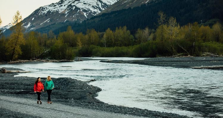 Two people walk alongside a rushing river beneath mountains.