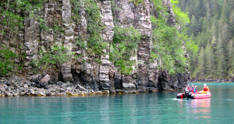 A group of scientists in a boat float alongside plant-covered cliffs.