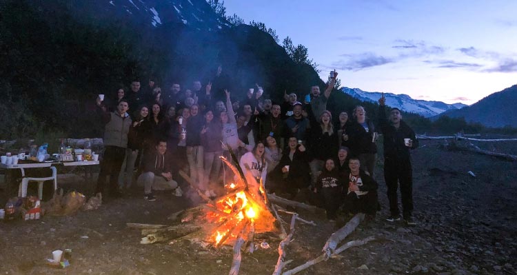 A crowd gathers around a bonfire on a rocky rivershore.