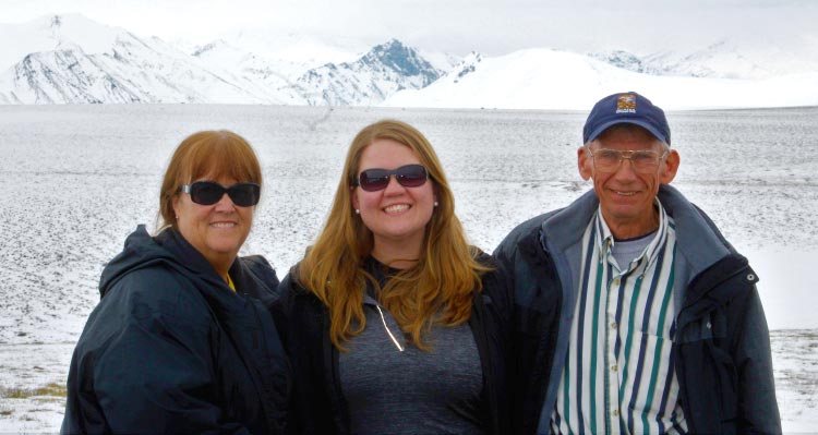 Three people stand before the sea with ice-covered mountains far behind them.