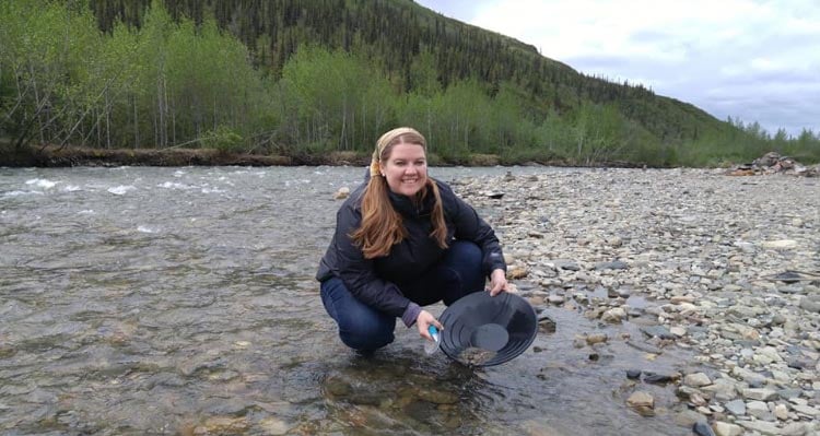 A woman crouches in a rocky river, panning for gold.