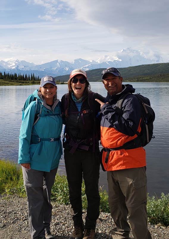 Three people stand and smile in front of a crisp lake in a tundra and mountain landscape.