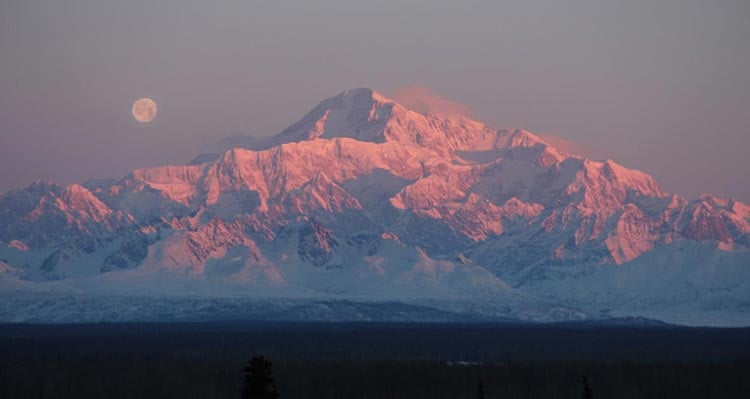 Denali, a tall mountain, covered in snow below a full moon.