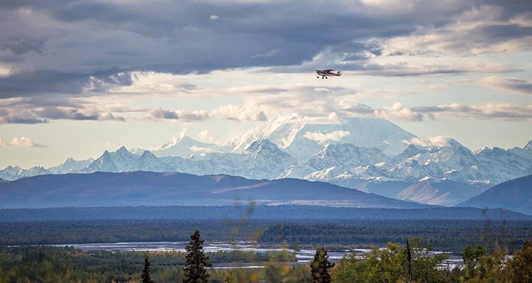 A small plane flies above an open Alaska landscape with Denali in the background.