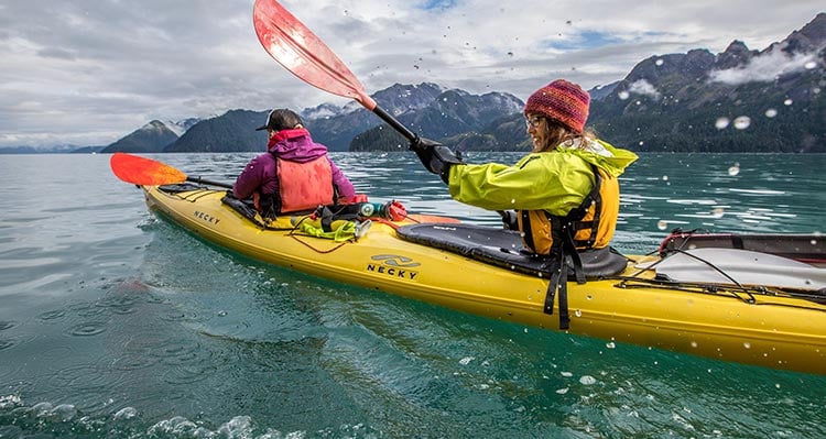 A pair of kayakers paddle on open water near green mountains.