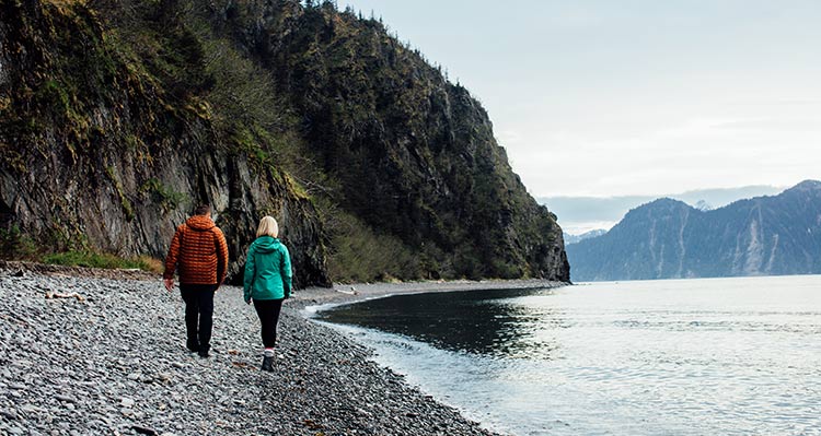 Two people walk along a rocky beach below a cliffside