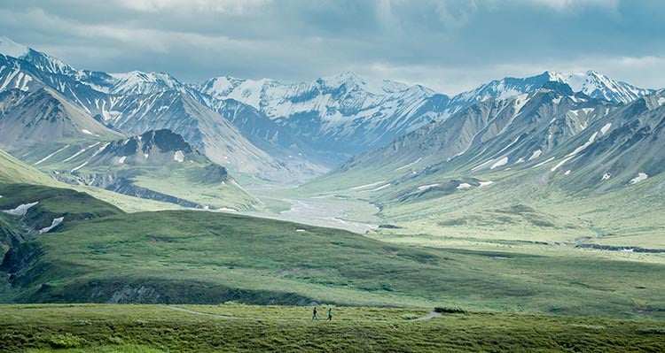 Two hikers walk along a lush landscape in a wide valley.