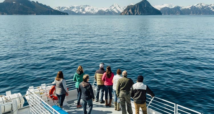 A small group of people stand at the edge of a boat railing looking towards green and snowy islands.