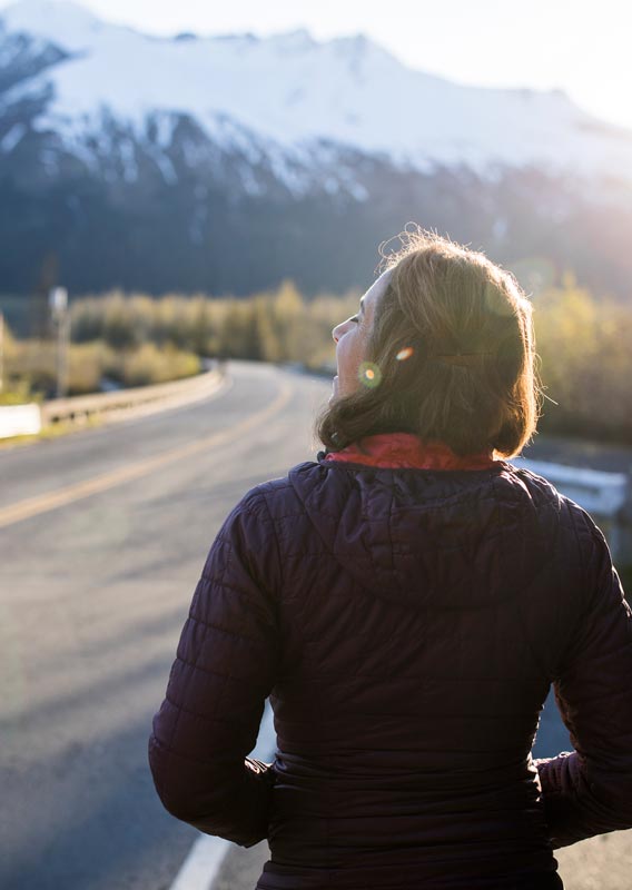 A woman looks down a sunny open road towards snow-covered mountains.