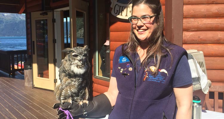 A woman holds an owl on her arm outside a wooden building.