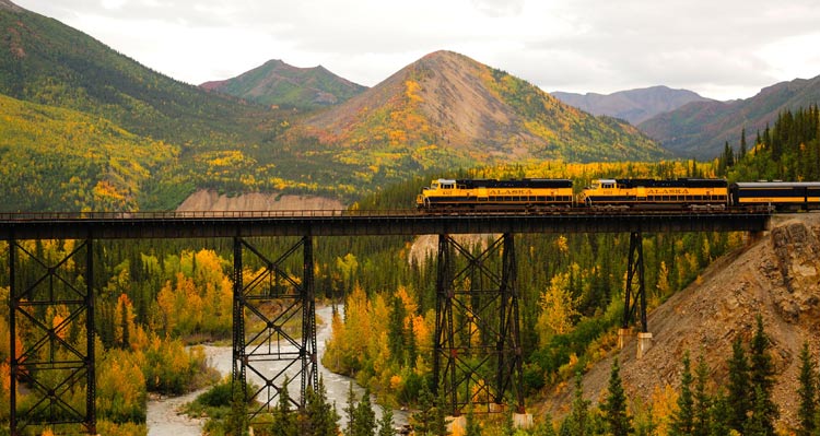 A train crosses a tall bridge over a river around forested mountains.