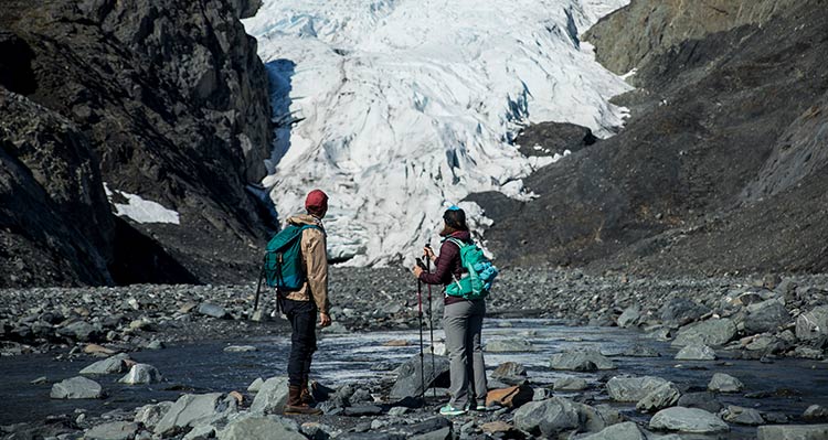 Two hikers stand on some rocks before a glacier.