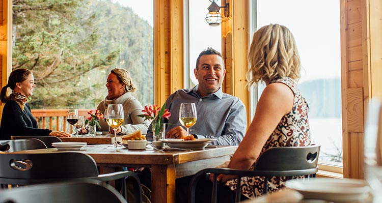 Two pairs of people sit at two tables eating in a wooden lodge with tall windows.