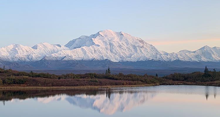 A landscape view of Denali, a tall mountain rising above other snow-covered mountains and a tundra landscape.