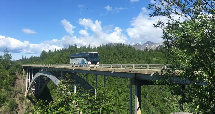 A motorcoach drives on a bridge between forests and mountains.