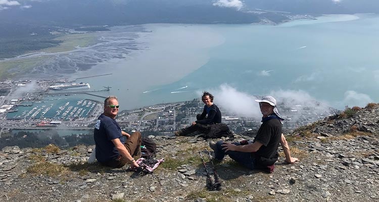 Three friends sit at a rocky outcrop overlooking the town of Seward and its harbor.