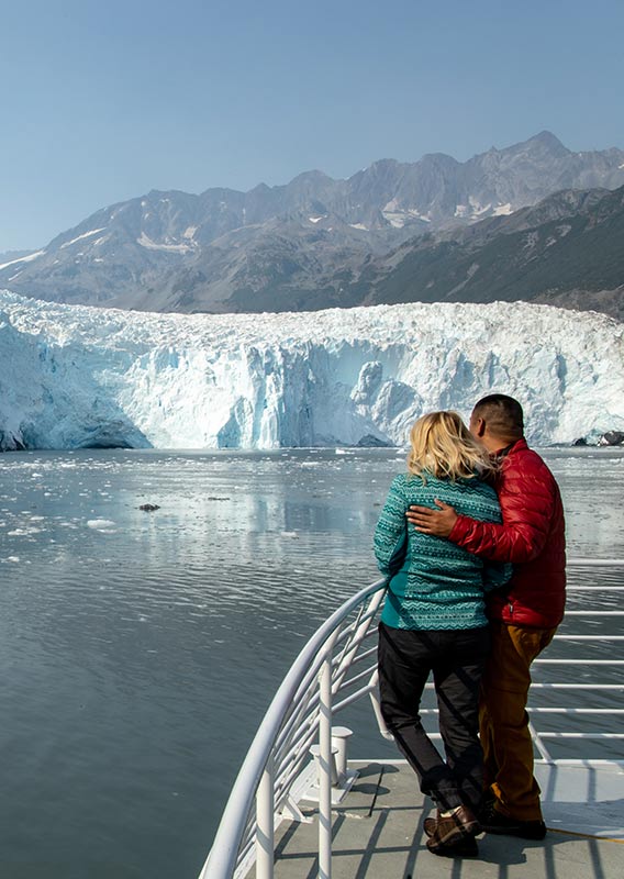 A couple at the helm of a tour boat views a glacier across waters of Kenai Fjords