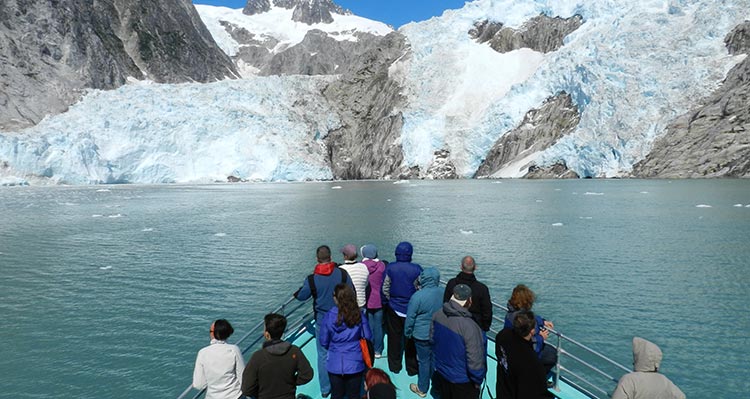 A group of people stand at the front of a boat approaching a glacier.