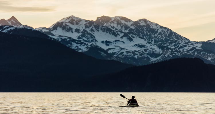 A sea kayaker paddles on the water with mountains ahead.