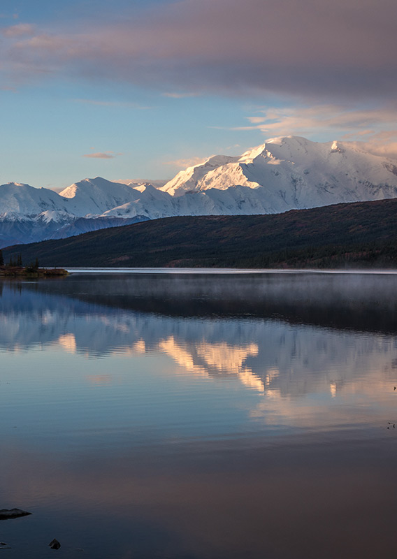 Snow covered Denali with sun shining on it's faces with a pristine lake at the base