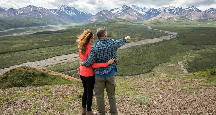 Two people stand looking out across a tundra landscape towards snow-dusted mountains.