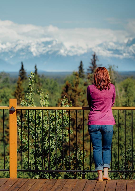 A person stands at a balcony fence looking towards a snowy mountain range.