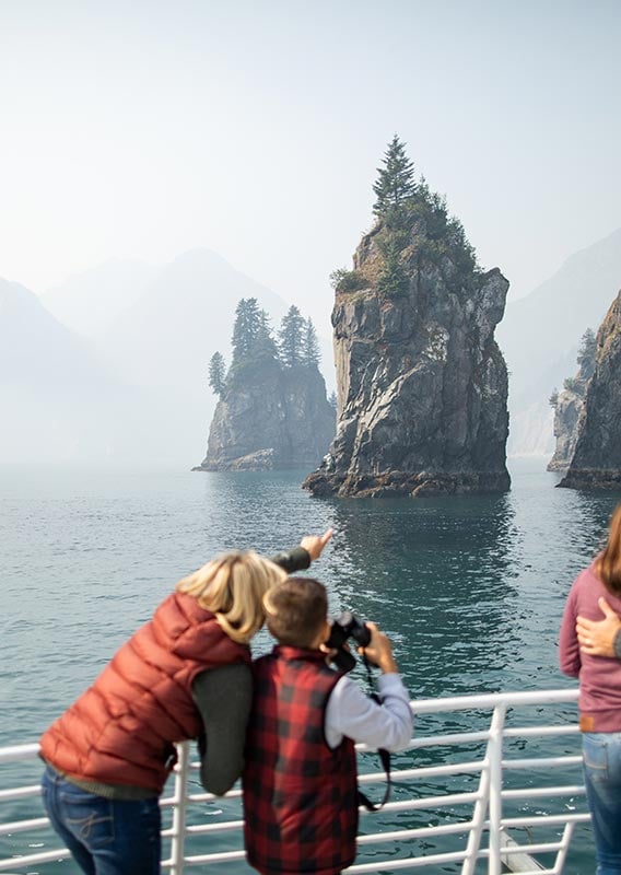 Groups of people stand at the railing of a boat looking towards rock pillars coming from the sea.