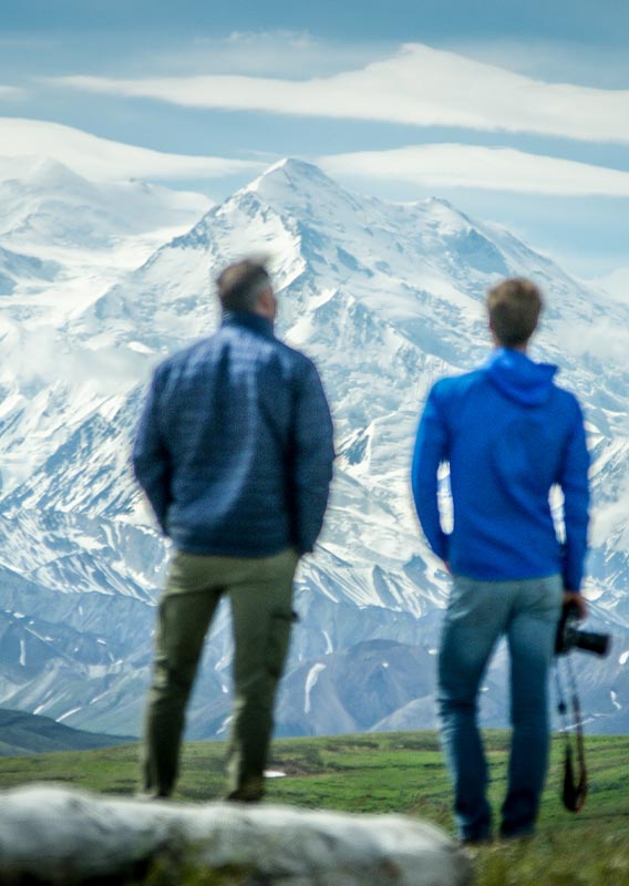 Two people stand at a viewpoint looking towards Denali, a tall snow-covered mountain.