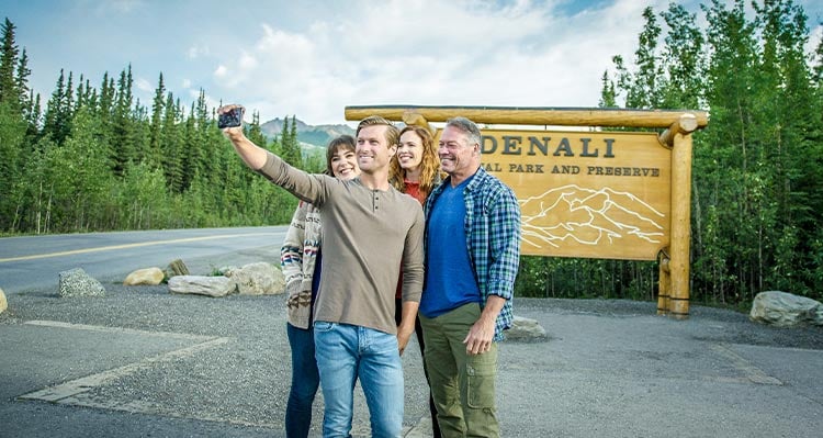 A group of people take a selfie in front of the Denali National Park sign.