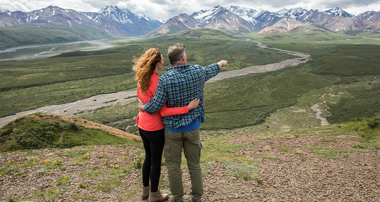 Two people look out from a viewpoint across a wide valley and mountains.