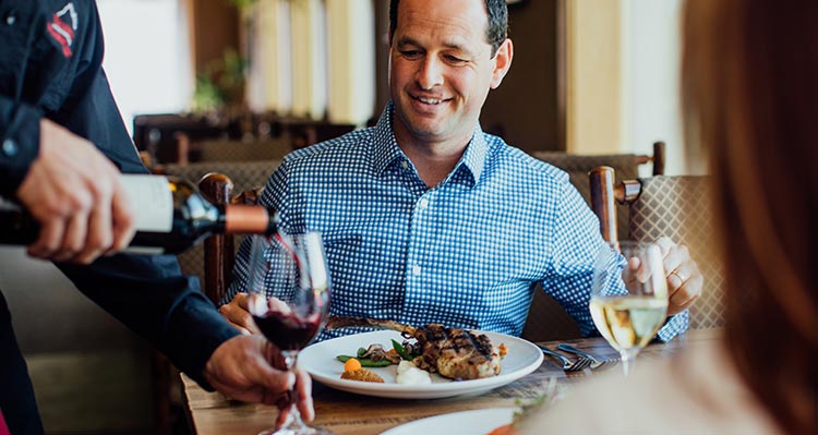 Two people sit to dinner while a waiter pours wine.