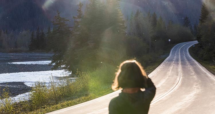 A person looks down an empty road near a rocky riverside.