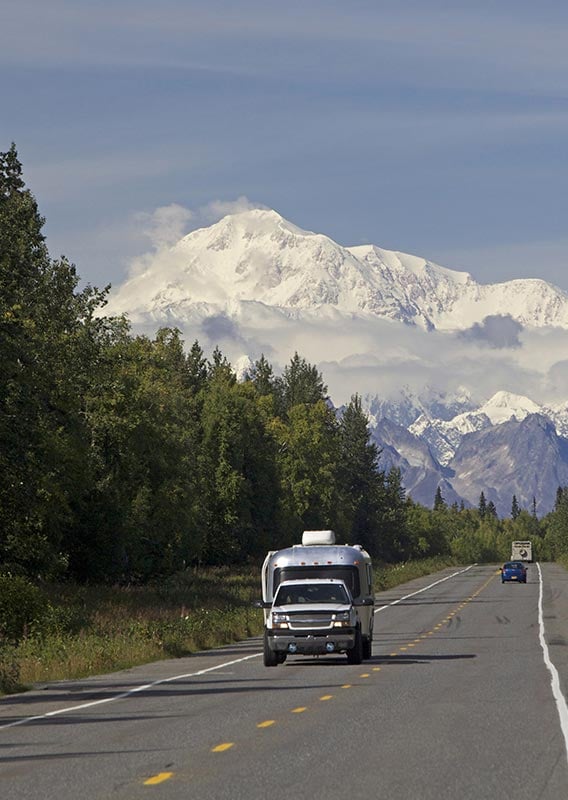 A truck pulling a camping trailer drives down a road with forests on both sides.