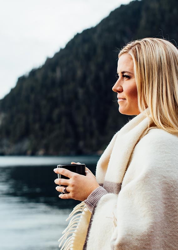 A woman stands on a seashore with a cup of coffee, wrapped in a blanket.